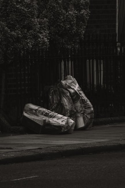 Two large black garbage bags made from durable plastic, filled with waste, are placed on the pavement beside a black metal fence with vertical slats. The bags appear to contain mixed rubbish, possibly household or garden waste. They are situated on a concrete sidewalk and partially leant against the fence, which runs along the edge of a residential or urban area. In the background, dense foliage from a tree or shrub partially obscures the upper section of the fence, with the scene illuminated by daylight, creating a clear, sharply defined contrast between the dark fence and the lighter street environment. The environment suggests an informal rubbish collection point, potentially indicating private waste disposal outside a property, relevant to services like independent rubbish clearance or alternative waste handling, as managed by companies such as Rubbish Clearance West Kensington.