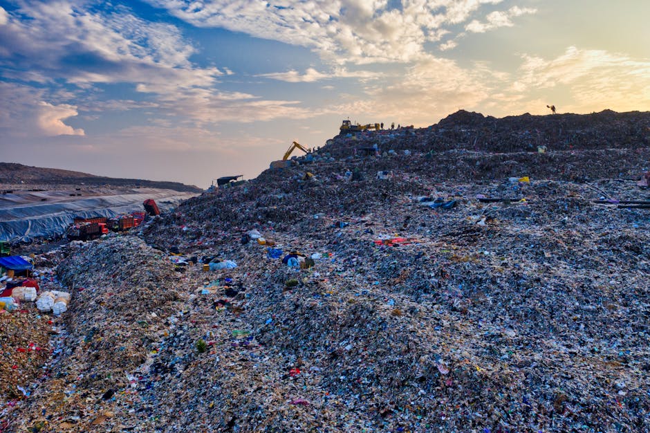 A large open landfill site filled with mounds of mixed waste materials including plastics, paper, textiles, and construction debris, all piled high across the landscape. The surface of the waste appears coarse and uneven, with visible textures of crushed, shredded, and compacted refuse. Brightly colored objects and packaging are scattered throughout, with some wrapped in black, white, or clear plastic coverings. In the background, heavy machinery such as excavators and dump trucks are positioned on the waste heaps, indicating active site operations. The sky overhead is partly cloudy with patches of sunlight casting a diffuse glow over the scene, enhancing the varied textures and colours of the waste. The environment surrounding the landfill is flat and expansive, with distant horizons and no significant vegetation. The scene emphasizes the scale of waste accumulation, potentially reflecting the need for alternative waste handling or rubbish clearance services, such as those provided by companies like rubbishclearancewestkensington.co.uk, to address bulky rubbish delays and private disposal challenges.