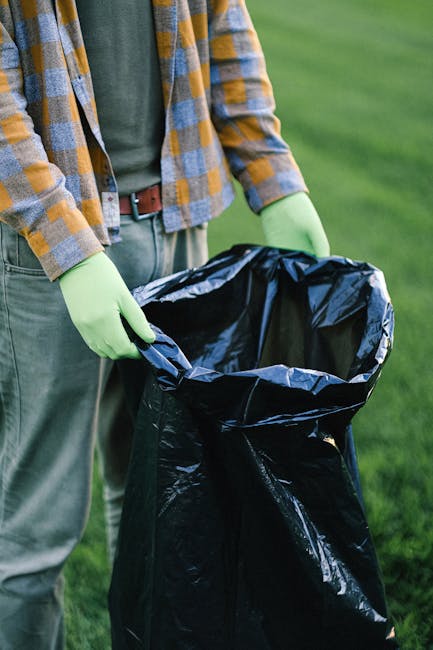 A person is standing outdoors on a grassy area, holding a large black plastic rubbish bag with both hands, which are gloved in bright green rubber gloves. The individual is dressed in casual clothing, including a checked shirt with shades of yellow, blue, and gray over a dark green t-shirt, and beige trousers secured with a brown belt. The background features a lush, green, well-maintained lawn, indicating the scene might be part of a garden or park setting. The black rubbish bag appears to be open, ready for rubbish collection, aligning with the context of waste management or waste disposal services. The scene emphasizes the manual handling of refuse, which could relate to private waste removal or on-site clearance carried out independently from local authority rubbish collection. The natural lighting enhances the clarity of textures, such as the glossy surface of the rubbish bag and the fabric of the clothing and gloves, illustrating a typical scene of rubbish disposal activity relevant for a professional waste management service reference.