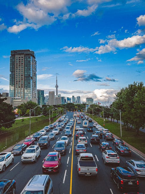 A busy multi-lane urban road filled with various vehicles including sedans, hatchbacks, and SUVs in different colours such as white, silver, black, and red, moving in both directions under a bright blue sky with scattered white clouds. To the left, a tall glass and steel office building with reflective windows dominates the skyline, while in the background, the cityscape extends with additional high-rise buildings and a prominent telecommunications tower with a spherical top. On the right side, green trees line the roadside, providing a natural contrast to the concrete and glass structures. The scene is well-lit with natural daylight, capturing the movement and density of city traffic, which may relate to the context of independent or private waste collection services in urban environments, such as those provided by rubbish clearance companies operating in West Kensington.