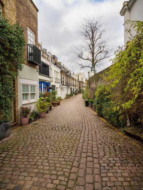 A narrow cobbled street in West Kensington lined with closely situated terraced houses, featuring small front gardens with potted plants on the left and a high brick wall covered in green foliage on the right. The residential buildings are mostly white or light-colored with some brick façades and small windows, with balconies or railings visible on upper floors. A leafless tree with thin branches stands at the end of the street, set against an overcast sky. The scene appears quiet and residential, with no visible vehicles or people, and the street's surface is composed of uneven, weathered cobblestones. The environment suggests an urban alleyway suitable for private rubbish removal or on-site clearance services, with the setting emphasizing the need for careful access and waste handling in a narrow, residential area, consistent with independent waste collection methods. The image captures a typical UK street scene emphasizing the importance of professional rubbish removal in such confined spaces, as supported by local waste management services.