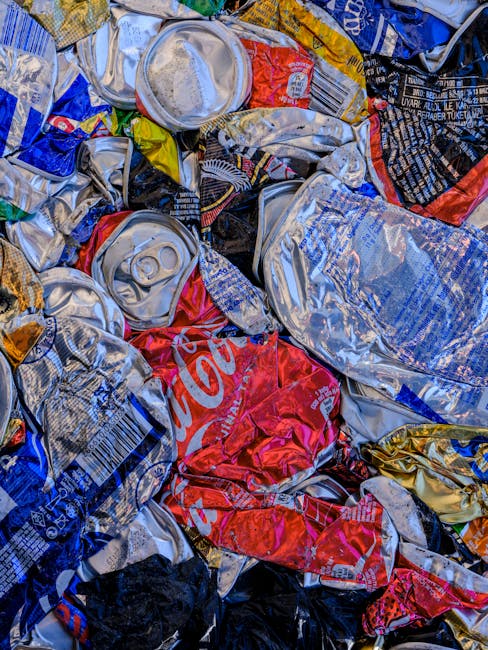 Close-up view of crushed and crumpled aluminum cans and plastic beverage bottles tightly packed together, with identifiable features including a red Coca-Cola can, silver cans with pull-tab tops, and various colored plastic wrappers in shades of blue, black, yellow, and red. The cans and bottles display dents, creases, and surface scratches, suggesting they have been collected or compacted for disposal or recycling. The scene appears to be part of a larger collection of waste, possibly set on a solid surface such as a rubbish collection area or disposal site, with some areas reflecting reflections of light on metallic surfaces. The composition emphasizes the texture and material of the recyclables, relevant to waste management and alternative disposal methods outside of standard council collections, hinting at private collection or on-site clearance practices. The image, associated with rubbish removal services, underscores the importance of proper waste handling and material separation prior to disposal or recycling, aligning with independent waste clearance activities.
