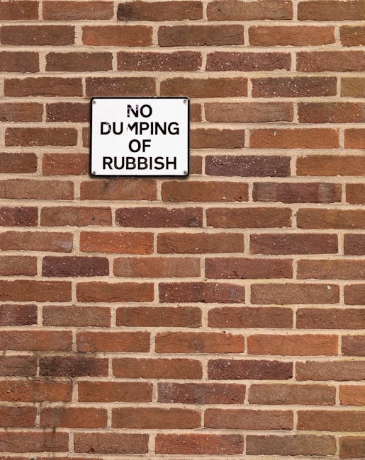 A rectangular black and white metal sign mounted on a red brick wall, displaying the message 'NO DUMPING OF RUBBISH' in bold uppercase letters. The bricks are uniformly laid with visible mortar joints, featuring a warm reddish-brown tone with some variations in shade. The sign is positioned slightly above the center of the image, with the brick wall extending evenly in all directions around it. The lighting is natural, casting subtle shadows to the lower left, and the overall scene suggests an outdoor environment, possibly a back alley or industrial area that could be subject to private rubbish collection or site clearance, aligning with the context of waste management and rubbish removal services.