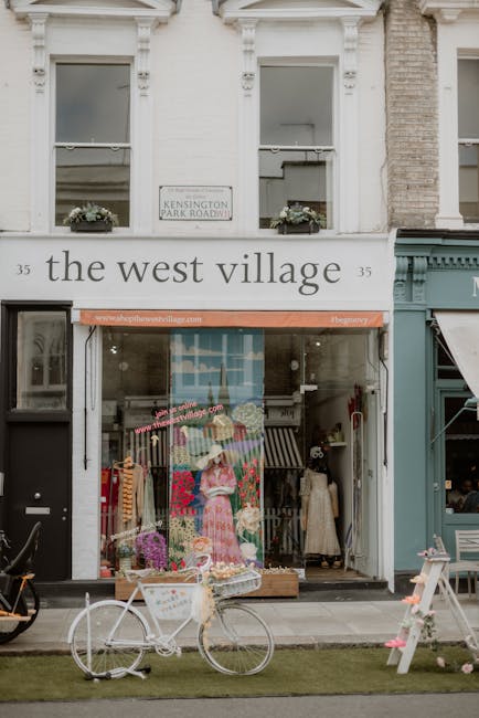 The image depicts the storefront of a retail shop named 'the west village' located at 35 Talgarth Road, West Kensington, with a white facade and large glass windows displaying colorful clothing and accessories inside. Above the entrance, there is a white sign indicating the location as 'Kensington Park Roadway' and a smaller sign on the storefront reads 'www.thelocalvillage.com'. In front of the shop, a vintage-style white bicycle with a rear basket is parked on a narrow, paved sidewalk, partially on a small patch of grass. Beside the bicycle, there is a white wooden A-frame sign, possibly advertising or providing information, and some outdoor display elements. The shop’s interior appears bright with clothing racks and mannequins visible through the large glass windows. The building has two upper-level windows with white frames, flower boxes, and decorative molding, contributing to its classic Victorian architectural style. The scene is well-lit with natural daylight, capturing an urban street scene in a commercial area that could relate to independent retail or local shopping precincts, typical for private waste collection or clearance services often used in such settings.