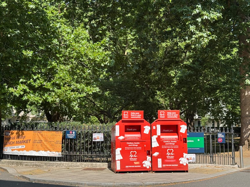 Two red charity collection boxes, designed for donating clothing, books, and other items, are positioned side by side on a paved sidewalk near a black metal fence. The boxes are made of metal with a matte finish, featuring white illustrations and text, including a heart logo and donation instructions. Each box has a slot at the top for inserting items, and the front panels display messages of gratitude and details about accepted donations. Behind the fence, there is a small green area with mature trees that have dense, vibrant green foliage, providing a shaded backdrop. To the left, part of an orange and white banner advertising a lunch market is visible, attached to the fence. The scene appears to be in an urban or suburban setting, with a bright and clear daytime atmosphere, natural sunlight illuminating the scene and casting shadows. The presence of the collection boxes suggests an alternative method for charity-based rubbish or item collection, supporting private disposal or donation services. This setting reflects a typical community space where waste clearance can include specific donation points managed by organisations or companies that handle rubbish and reuse efforts outside of local authority collection.