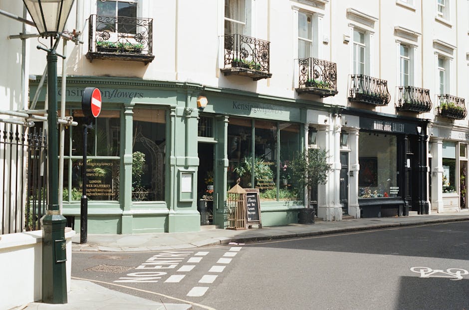 The image depicts a quiet street scene featuring a row of white-painted, multi-story commercial and residential buildings with black wrought-iron balconies adorned with flower boxes, some containing greenery. In the foreground, a small business storefront painted in pastel green has large display windows showcasing plants and flowers inside, with a chalkboard sign placed on the sidewalk outside advertising floral arrangements. To the right of the shop, another storefront painted in black also has large windows with displayed items. The street surface is paved, with white dashed lines indicating a designated bicycle lane, and a pedestrian crossing marked on the road. A tall street lamp with a classic design stands on the left side of the roadway, and a no-entry traffic sign attached to a pole is visible near the corner. The scene appears to be captured during daylight hours with natural lighting, suggesting a calm urban environment. This setting reflects a typical location where private waste collection or on-site clearance may be relevant, and it is aligned with professional rubbish removal services operating within residential and commercial areas in West Kensington.