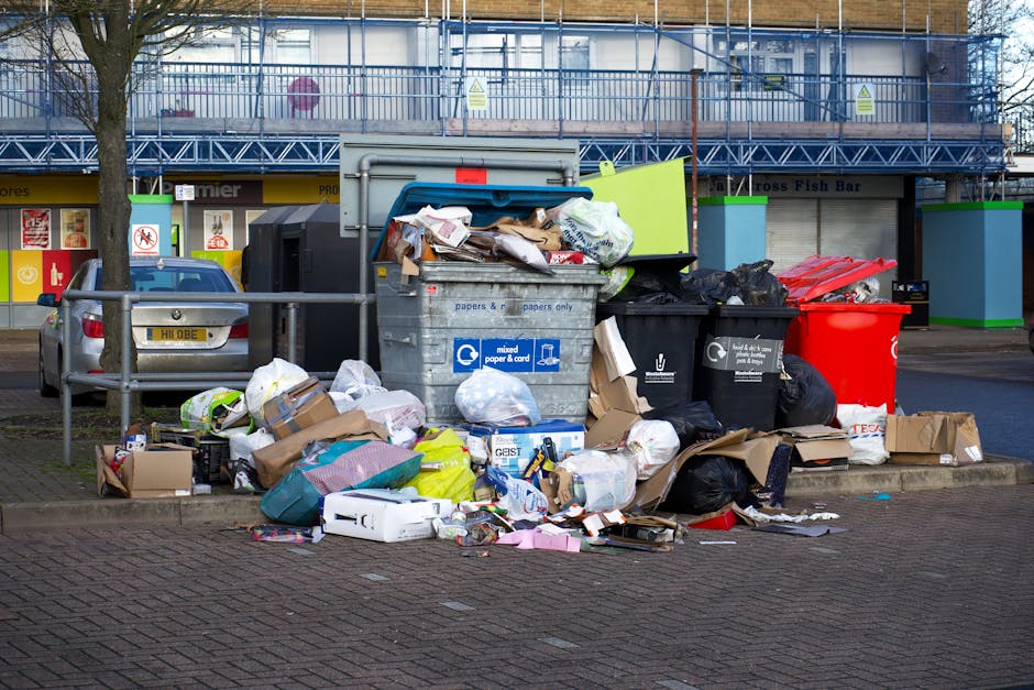 A large collection of mixed waste, including cardboard boxes, paper bags, plastic bottles, black and red plastic trash bins, and loose packaging materials, piled up on a paved area in front of a retail or commercial building. The waste appears to be a combination of recyclables and general rubbish, with some items spilling out of open bins and onto the surrounding pavement. To the left, a parked grey hatchback car is partially visible, positioned behind a metallic railing that separates the waste area from the pedestrian pathway. In the background, a multi-storey structure with scaffolding and blue safety netting indicates ongoing construction or maintenance work, and storefronts with signage are visible at street level. The scene is outdoors, with natural daylight illuminating the area, highlighting the disorganized nature of the waste and the environment's urban setting, which is typical of private rubbish collection points or on-site clearance areas managed by waste removal services such as those offered by Rubbish Clearance West Kensington.